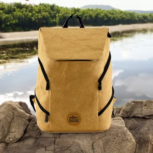 A yellow kraft paper laptop backpack with zippers and a logo, set against a scenic outdoor backdrop.