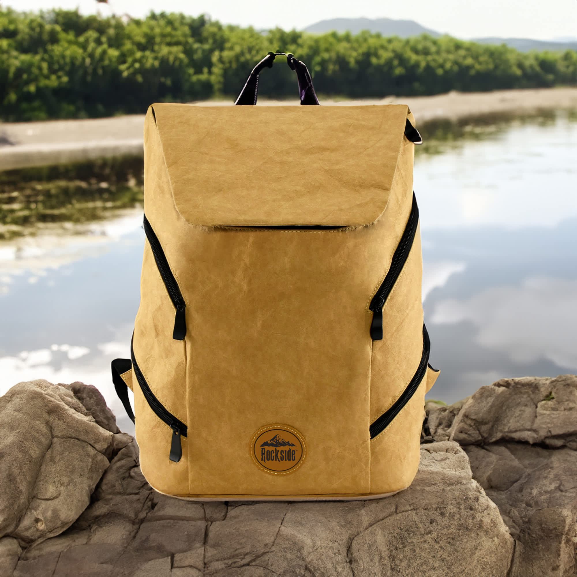 A yellow kraft paper laptop backpack with zippers and a logo, set against a scenic outdoor backdrop.