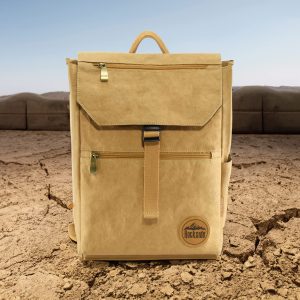 A tan kraft paper laptop backpack with a front pocket and branded logo, set against a dry, rugged landscape.
