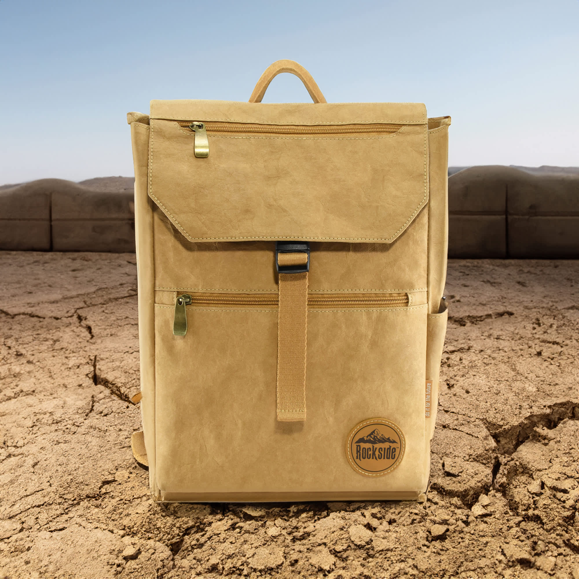 A tan kraft paper laptop backpack with a front pocket and branded logo, set against a dry, rugged landscape.