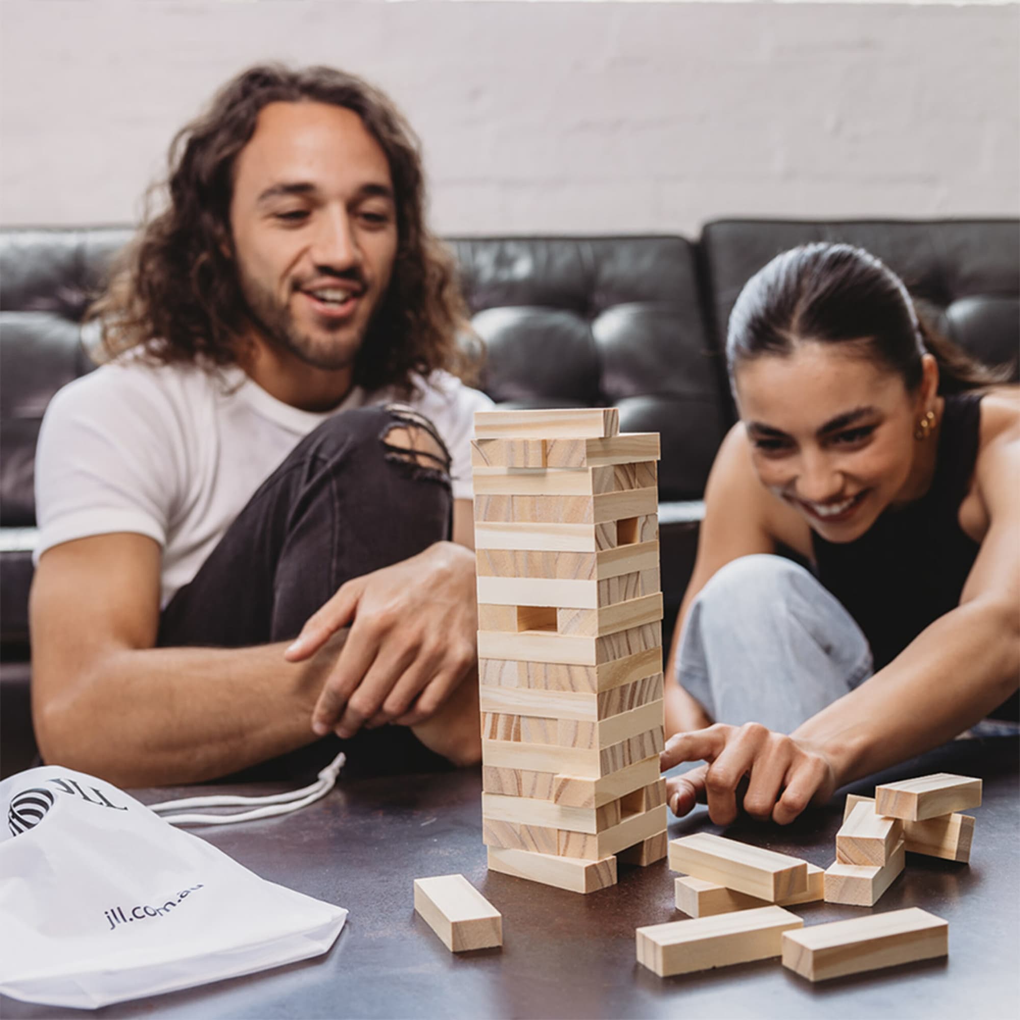 A wooden stacking puzzle set in a pouch, featuring light-coloured blocks arranged in a tower, with two people playing.