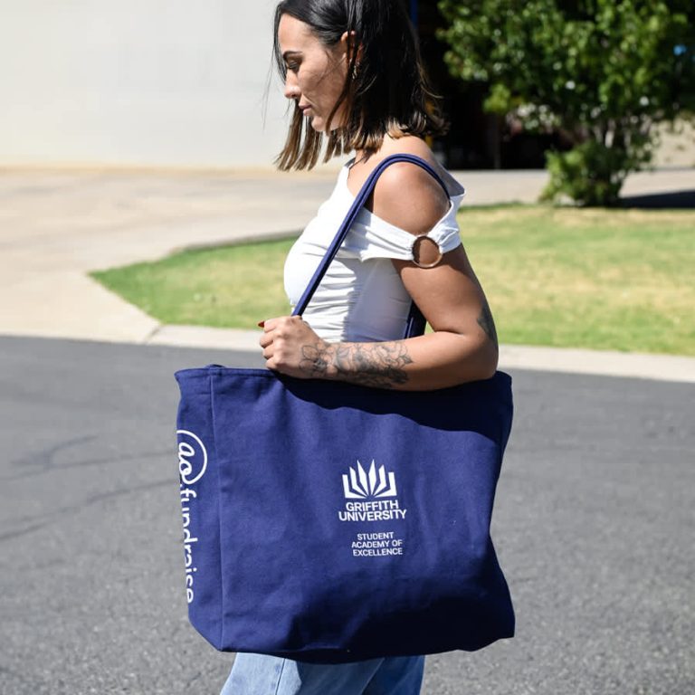 A navy tote bag with a logo, featuring sturdy handles and a spacious design, carried by a person.