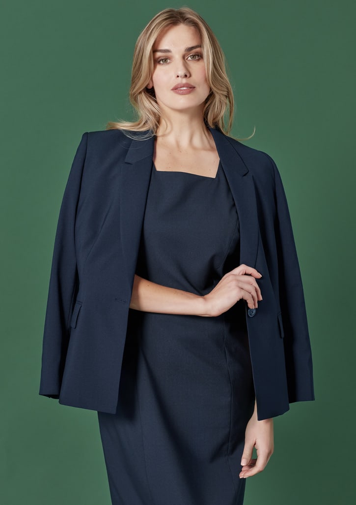 A woman wearing a fitted black blazer over a matching dress, set against a green background. The image features a logo.
