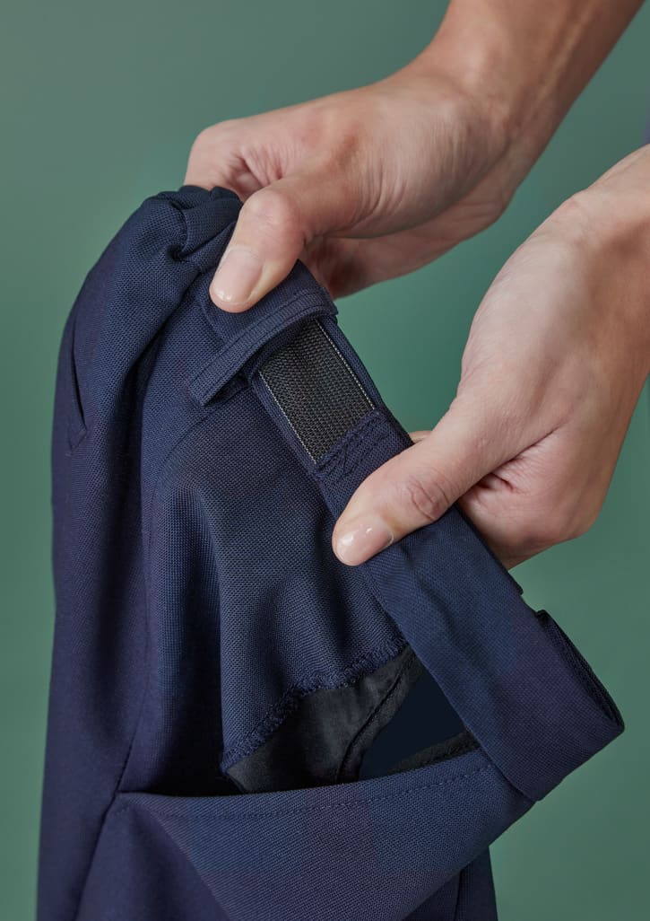 A close-up of hands adjusting the waistband of navy tapered pants featuring an adjustable strap and a logo.