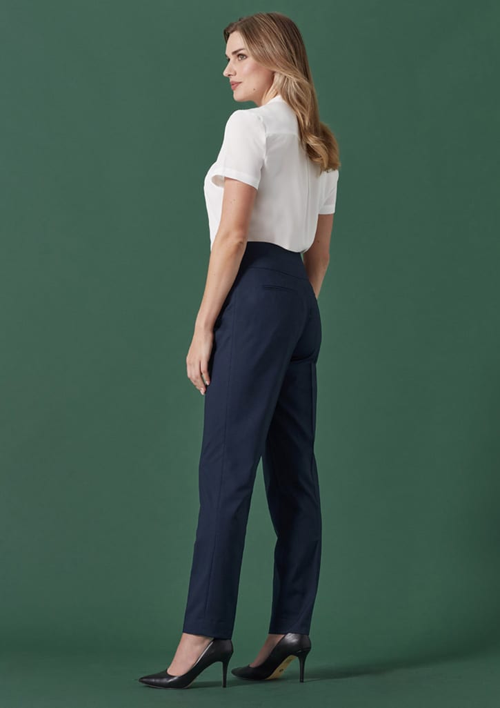 A woman stands sideways wearing mid-waist slim leg pants in navy, paired with a white shirt and heels against a green background.