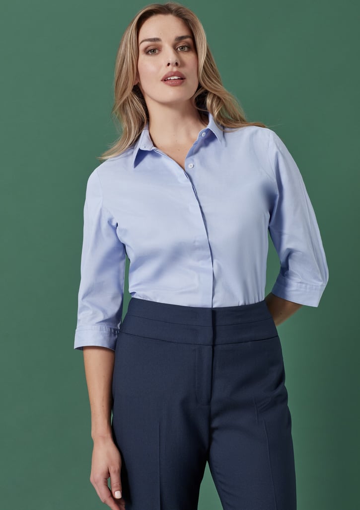 A woman wearing a light blue shirt and dark blue mid-waist slim leg pants against a green background.