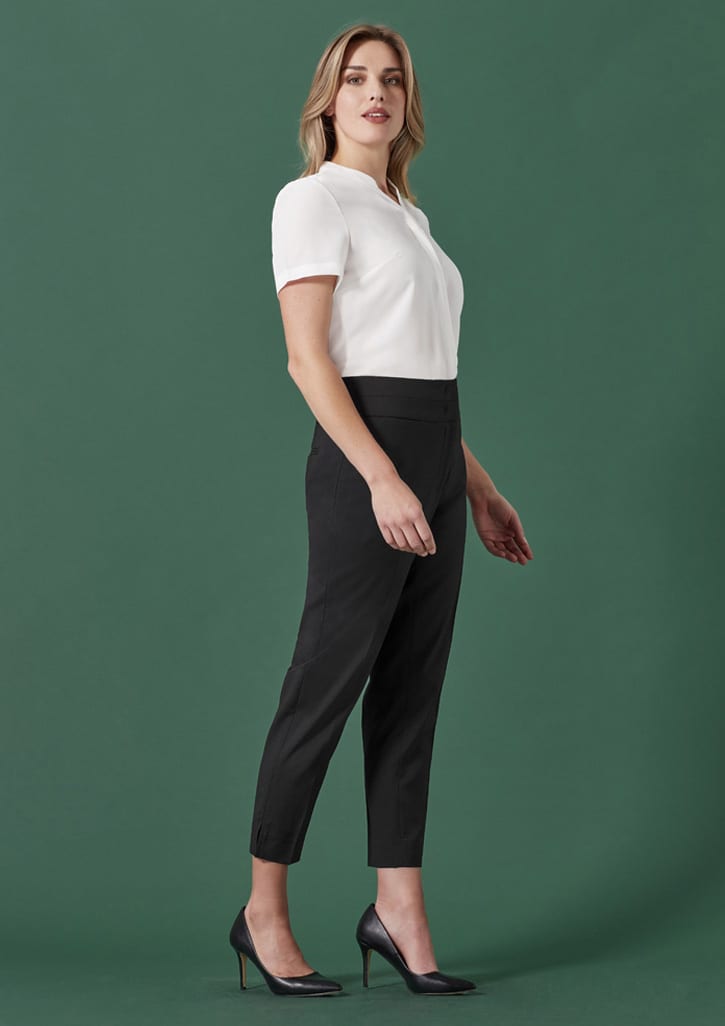 A woman stands in profile, wearing a white top and black mid-waist slim leg pants against a green background.