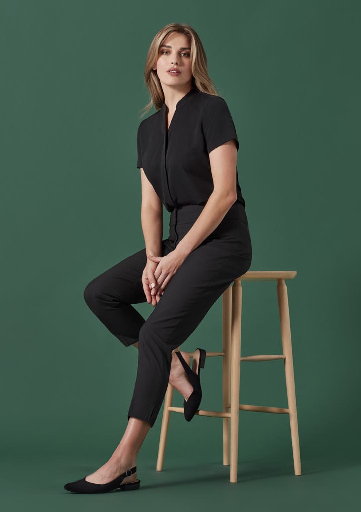 A woman in black mid-waist slim leg pants sits on a wooden stool against a green background.