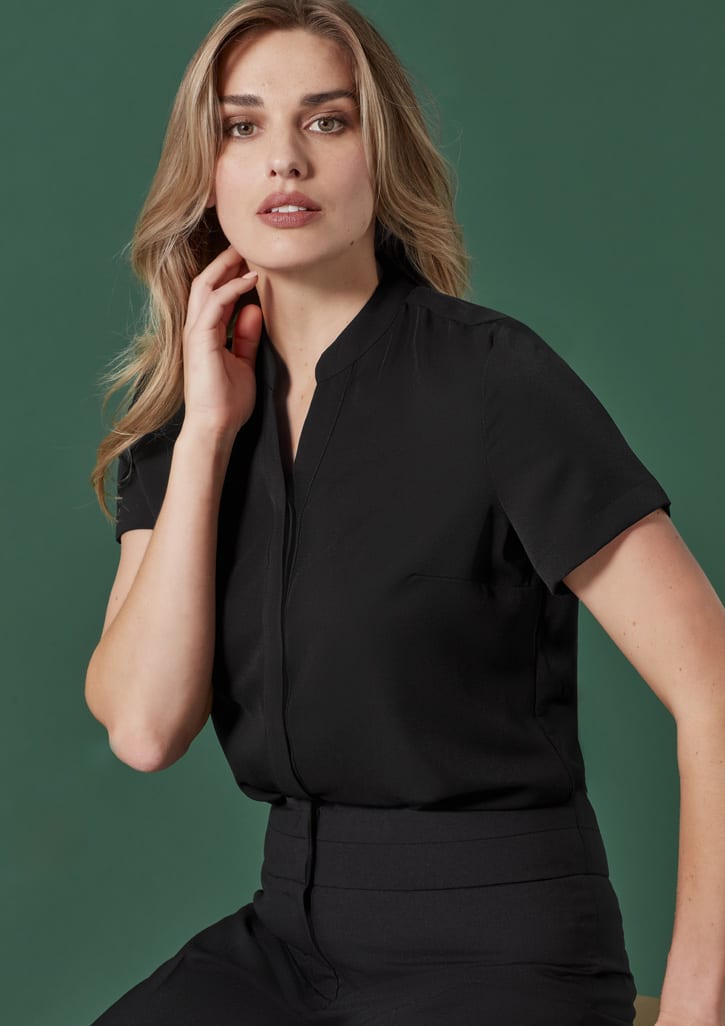 A model poses wearing a black short-sleeve blouse with a sleek collar against a green background.