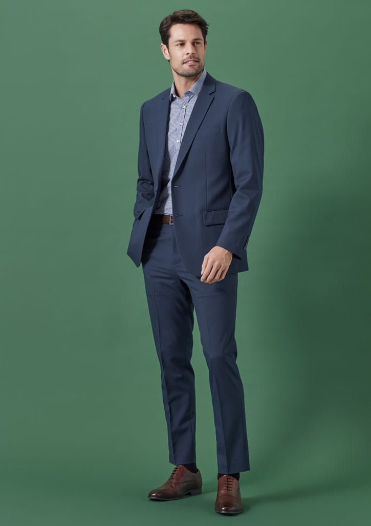 A man wearing a navy blue suit with a patterned shirt and brown shoes, standing against a green background.