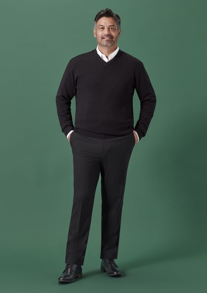 A man wearing a black sweater and black slim fit flat front pants stands against a green background.