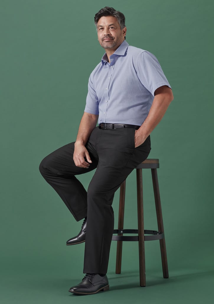 Men's straight leg pants in black, worn by a model seated on a stool. The model is dressed in a blue shirt and black shoes.