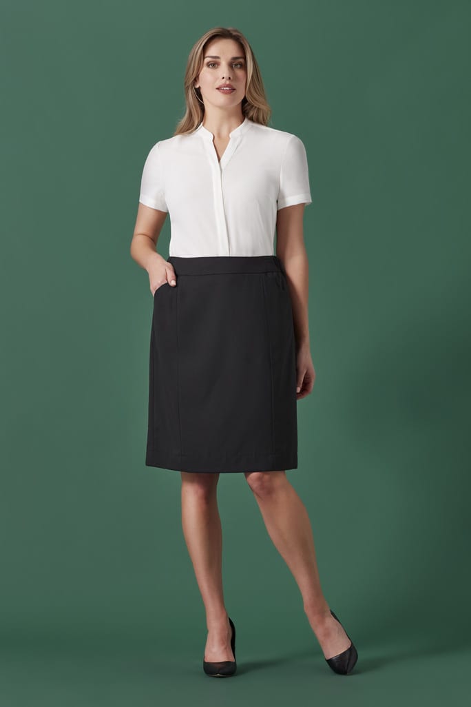 A woman in a white blouse and black functional pencil skirt stands against a green background. The skirt has pockets.