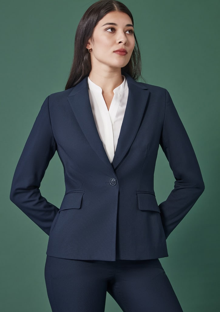 A navy single-button mid-length jacket with front pockets, styled with a white blouse. The model stands confidently against a green backdrop.