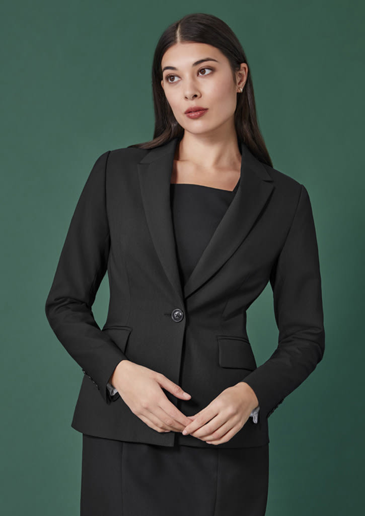 A women's mid-length blazer in black with a single button and two front pockets, modelled against a green background.