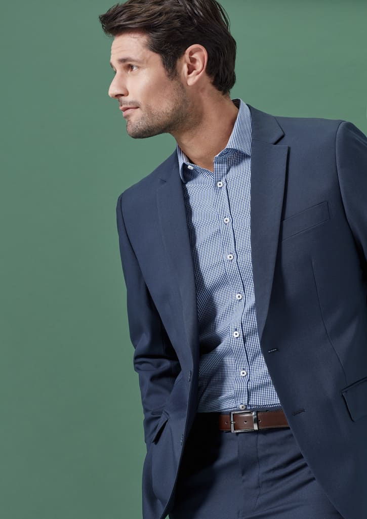 A man in a navy blue jacket over a striped shirt, standing against a green background. The jacket features a branding logo.