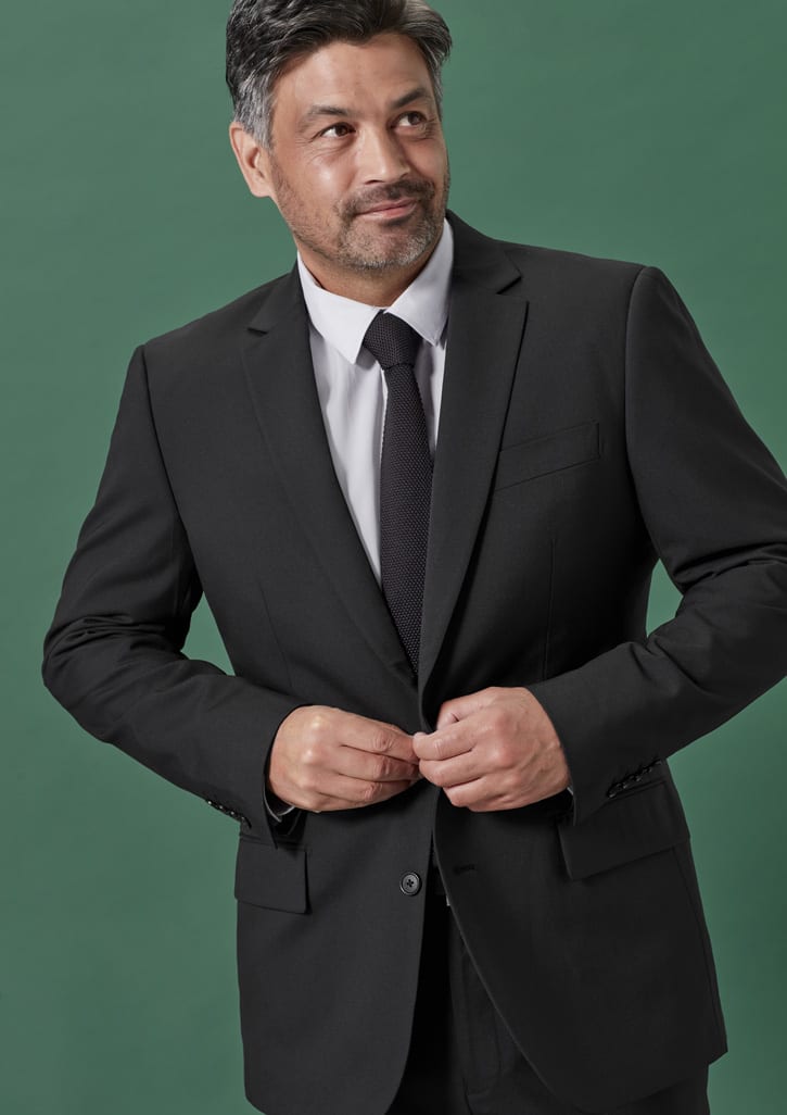 A man in a black suit jacket and tie, adjusting the buttons on his jacket against a green background.
