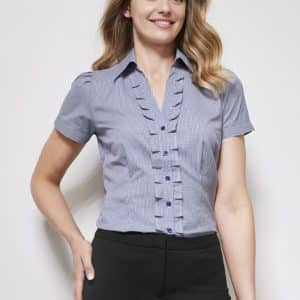 A short-sleeve button-up shirt in blue with ruffle detailing, worn by a smiling woman against a plain background.