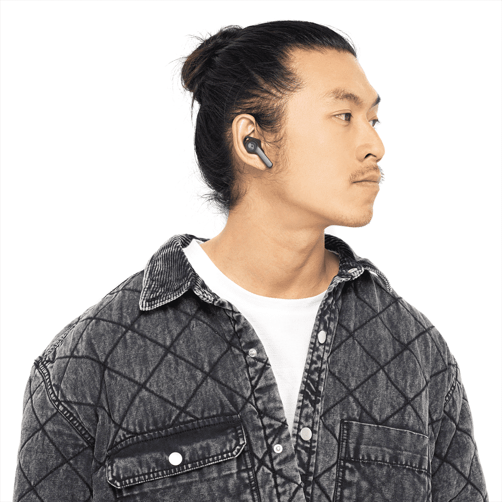 A person wearing black Skullcandy Indy EVO True Wireless earbuds, with a grey quilted shirt and a neutral background.