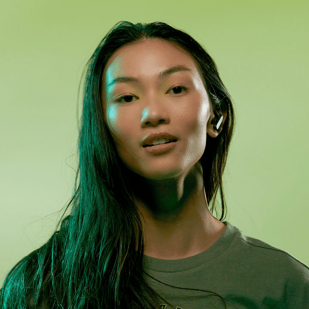 A model with long hair wears wireless earbuds against a green background. The design features a sleek, modern style.