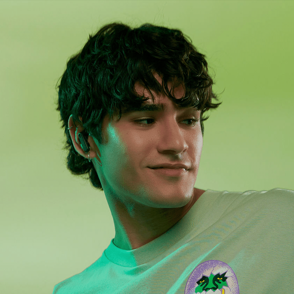 A young man with dark, wavy hair wears black earbuds against a green background. The product features a logo.