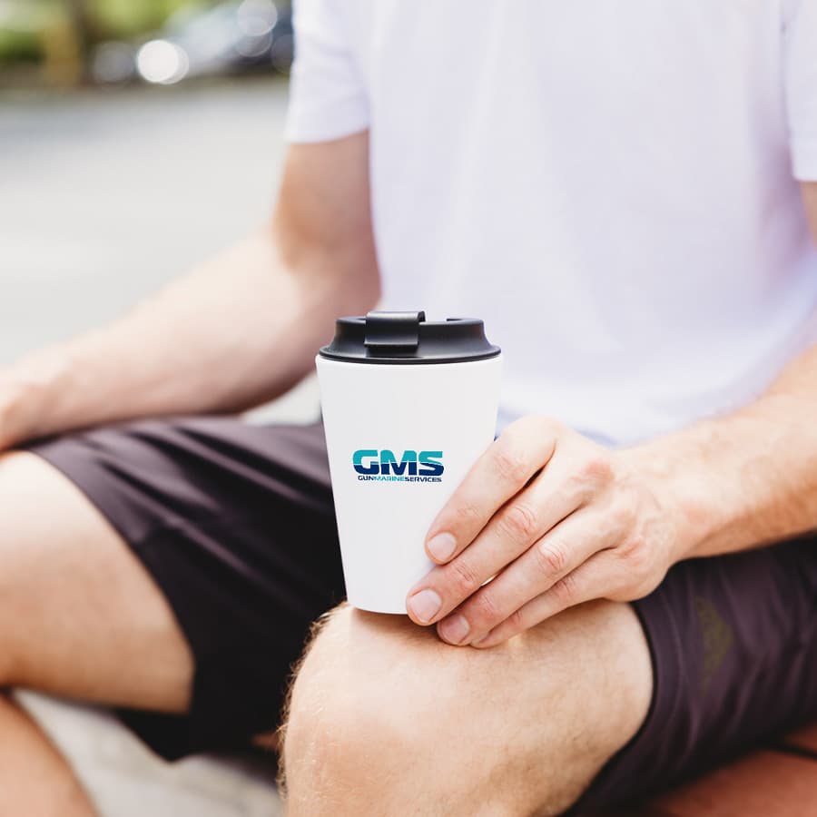 A coffee mug in white with a black lid, featuring a logo, held by a person sitting outdoors.