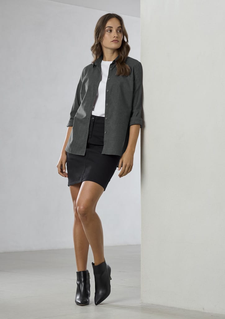 A woman wearing a grey button-up long sleeve shirt over a white t-shirt and a black skirt, paired with black ankle boots.