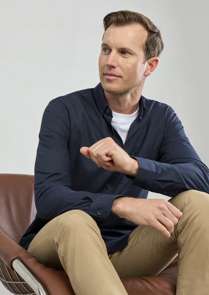 A man in a navy long sleeve button-up shirt sits casually on a chair, wearing light-coloured trousers.