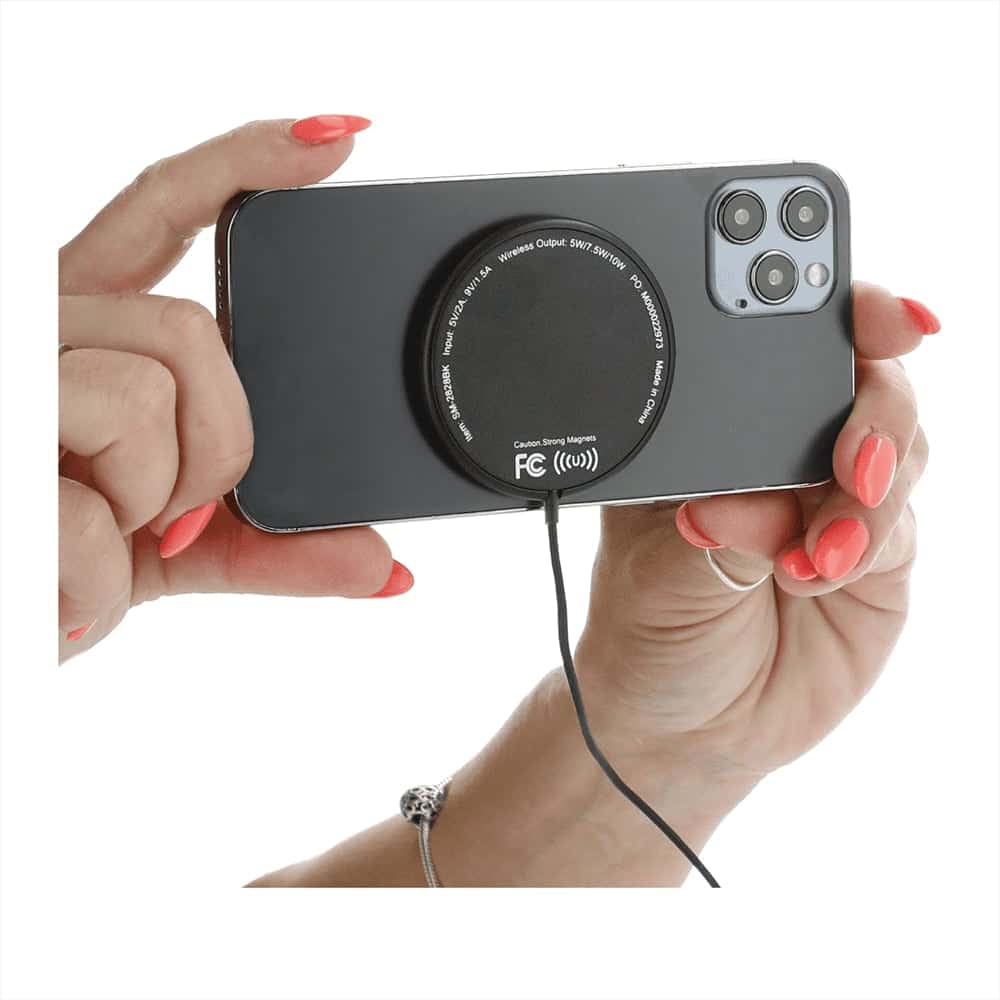 A black wireless charging pad is attached to the back of a smartphone held with a hand featuring pink nails.