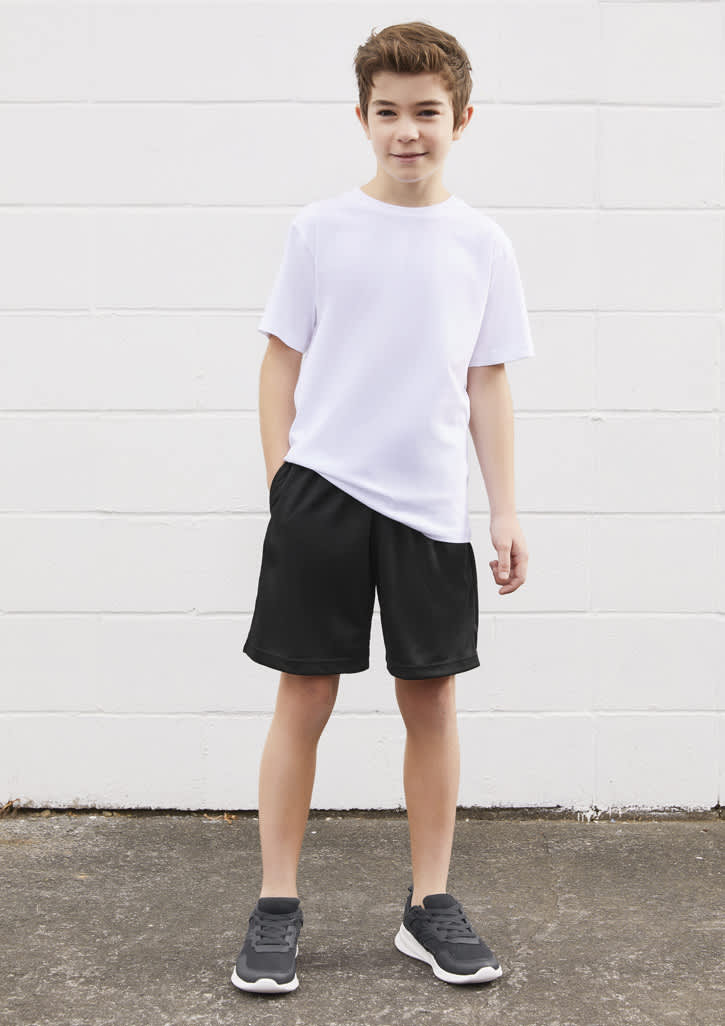 A boy wearing a white t-shirt and black sports shorts, standing against a white wall. The outfit includes footwear.
