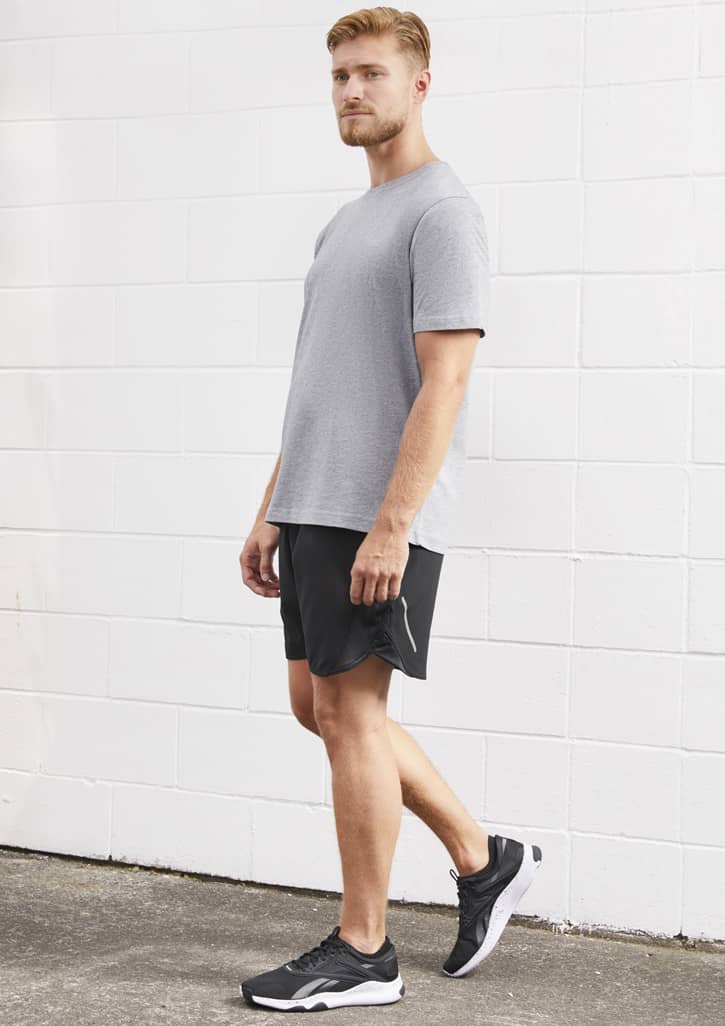 A man stands wearing grey sports shorts and a matching grey t-shirt, with black athletic shoes. The backdrop is white.