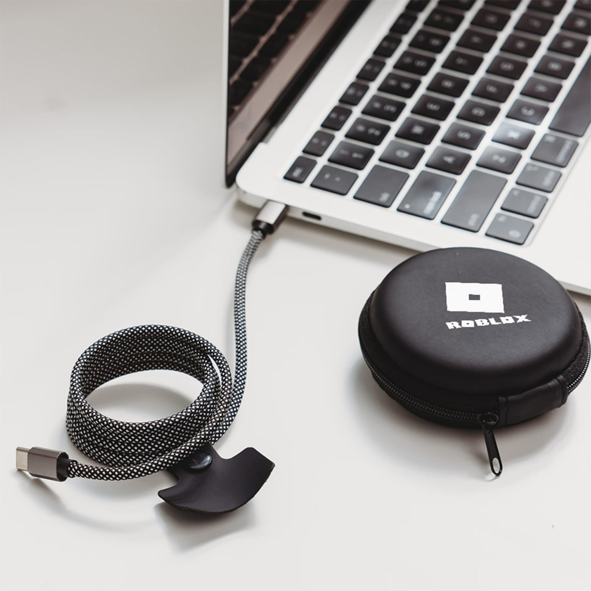 A charging cable in black and grey, coiled next to a round black case with a logo, connected to a laptop.