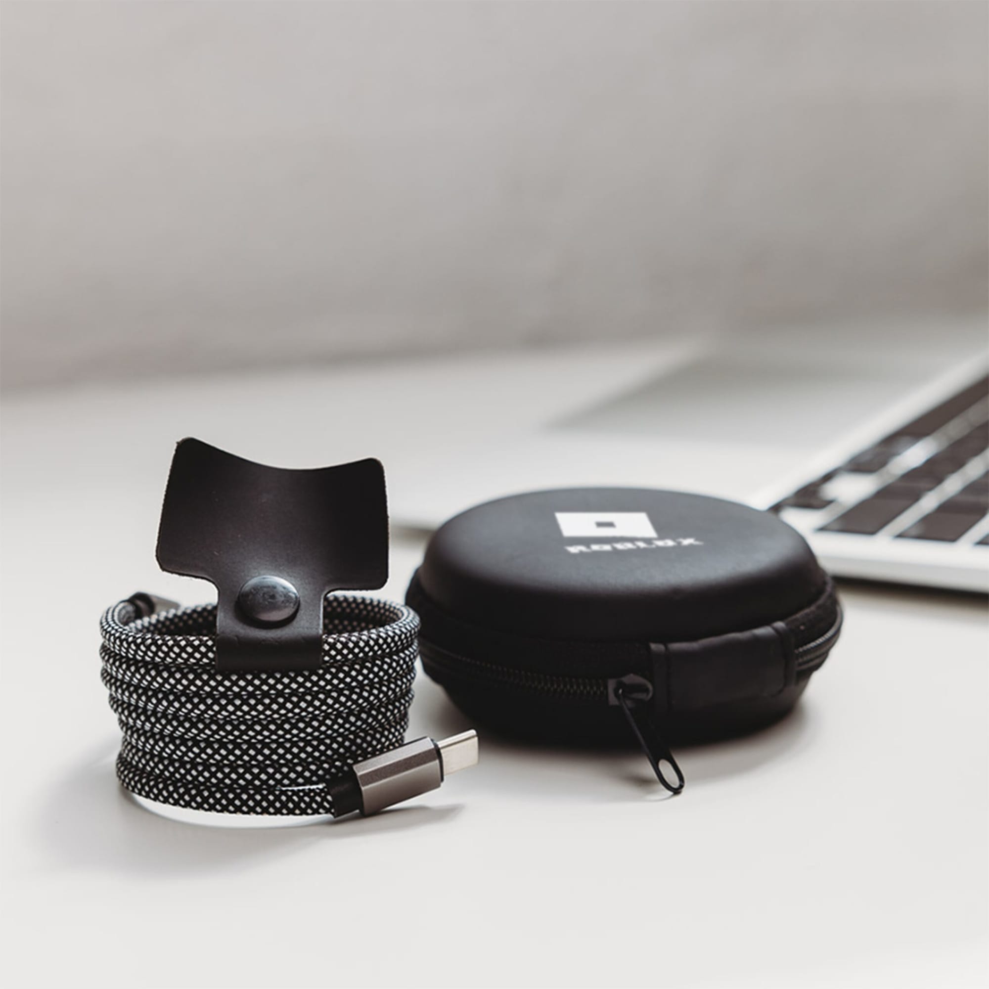 A charging cable in black and white fabric with a snap closure, packaged in a circular black case with a logo.