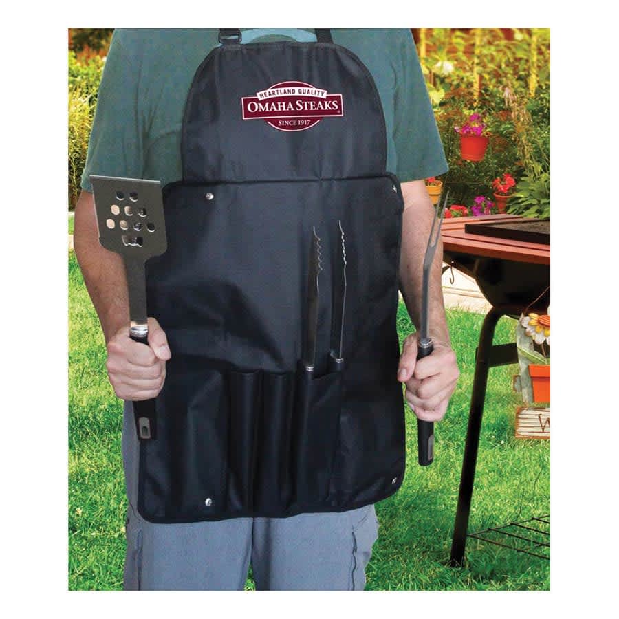 A man wearing a black apron holds a spatula and tongs, with a grill in the background. The apron features a logo.
