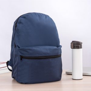 A navy blue backpack with a front zip pocket beside a white drink bottle on a desk.