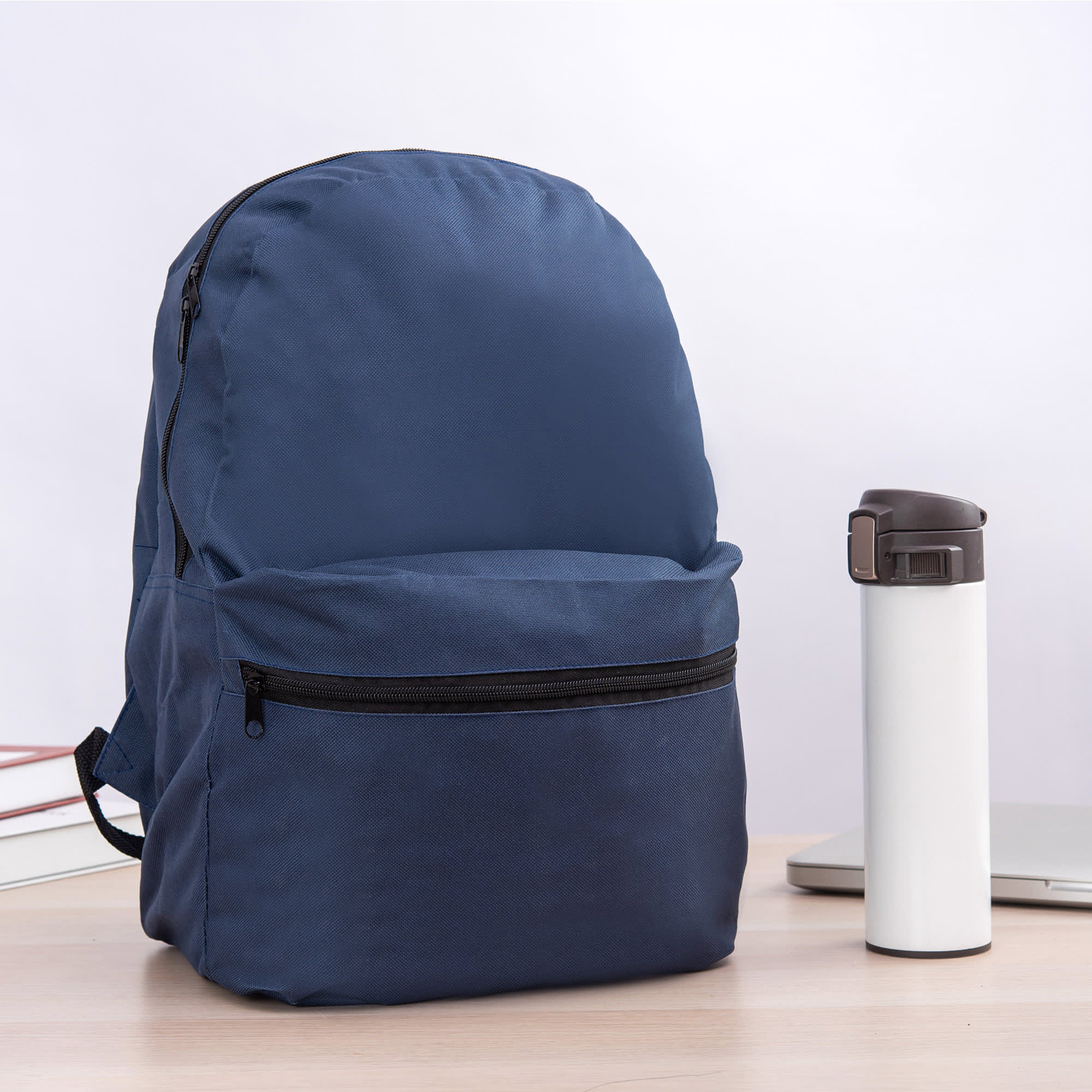 A navy blue backpack with a front zip pocket beside a white drink bottle on a desk.