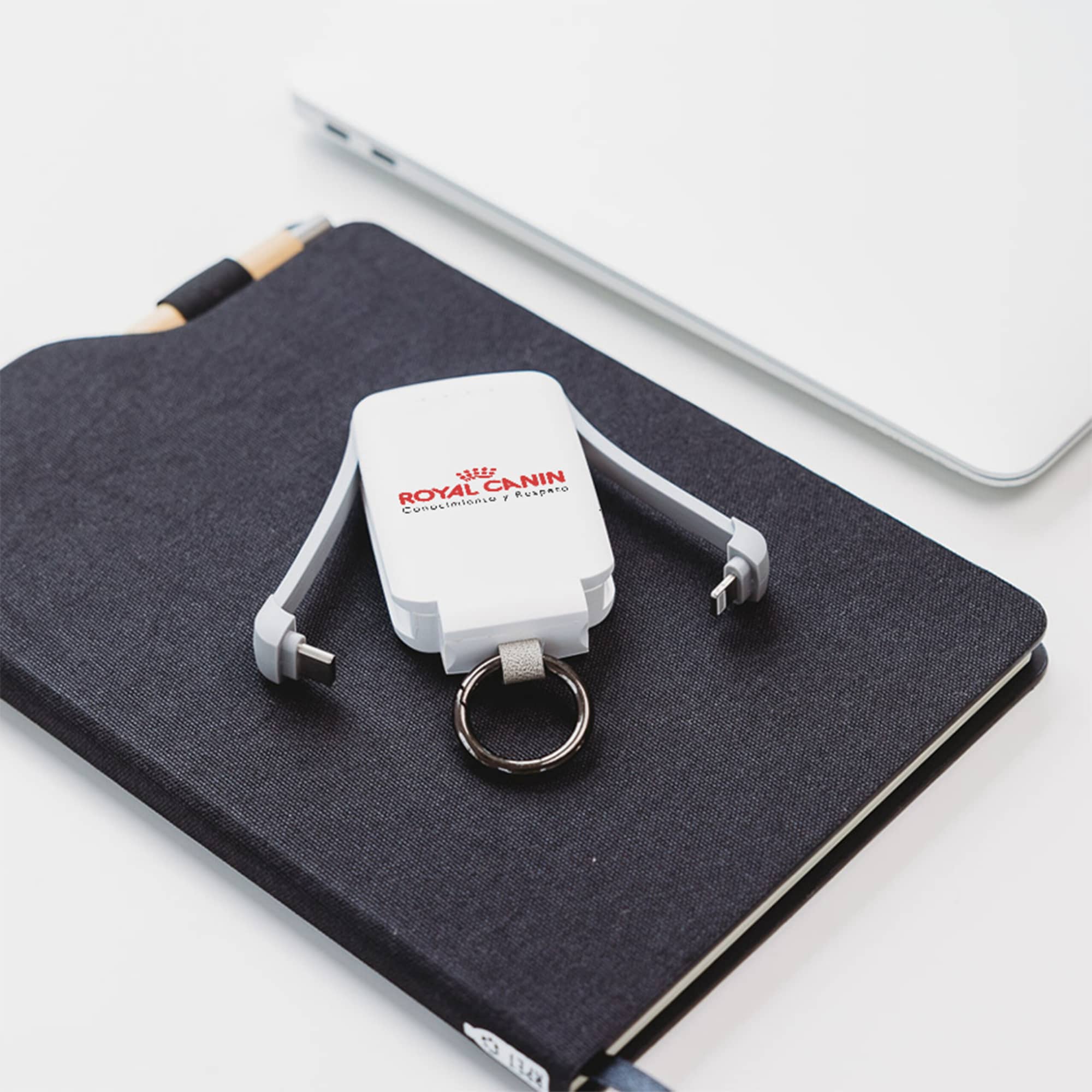 A white mini power bank with a keyring, placed on a dark notepad beside a silver laptop. It has a logo on it.