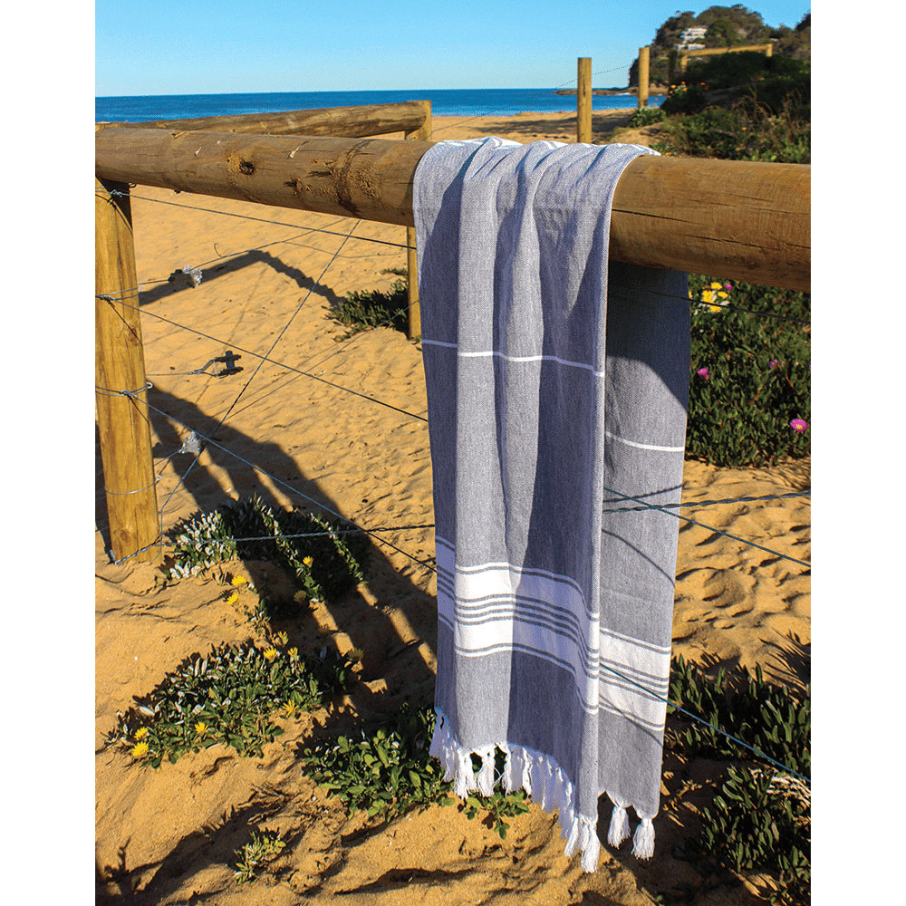 A grey and white striped Turkish towel draped over a wooden fence on a sandy beach, with ocean in the background.
