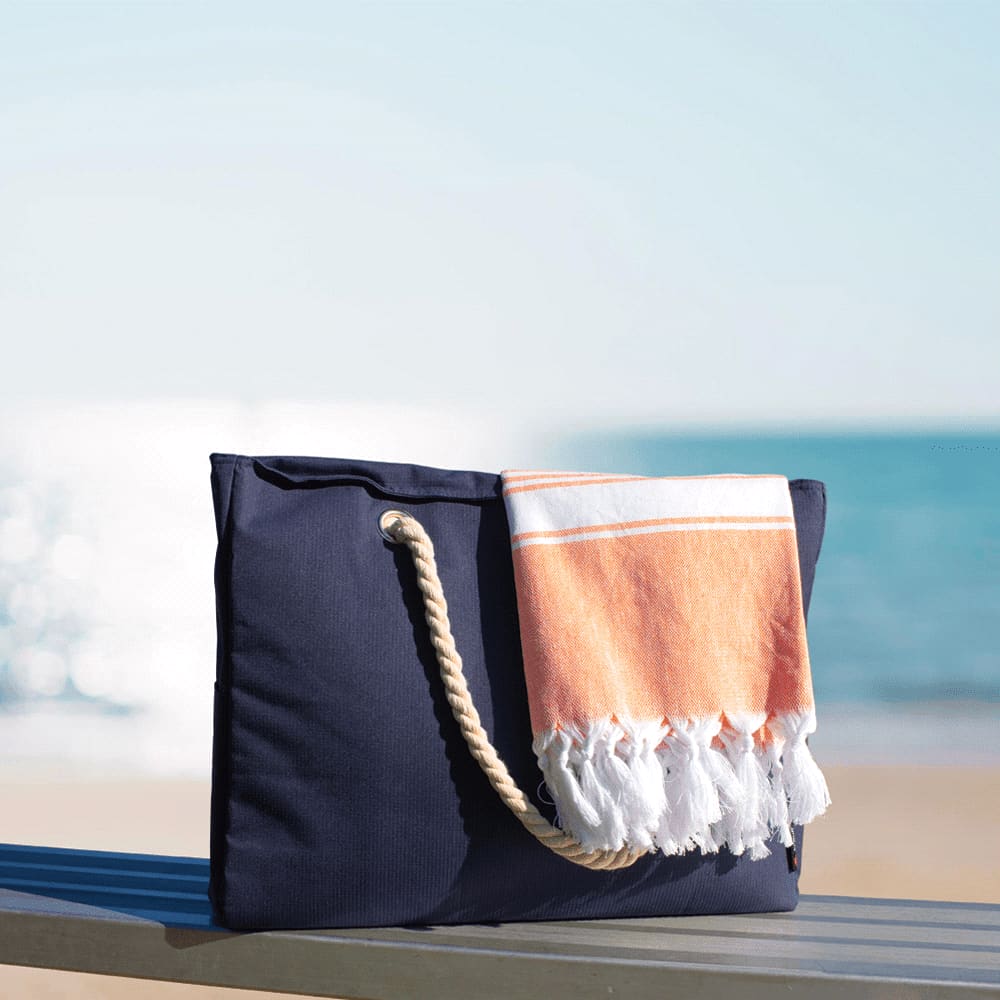 A navy waterproof beach bag with a woven handle and a folded orange and white towel draped over it.