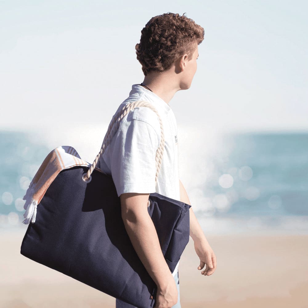A waterproof navy beach bag with rope handles, carried by a person against a coastal background.