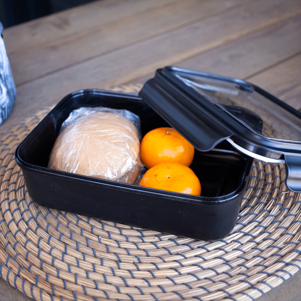 A black reusable container with a transparent lid, featuring a wrapped sandwich and two oranges inside.