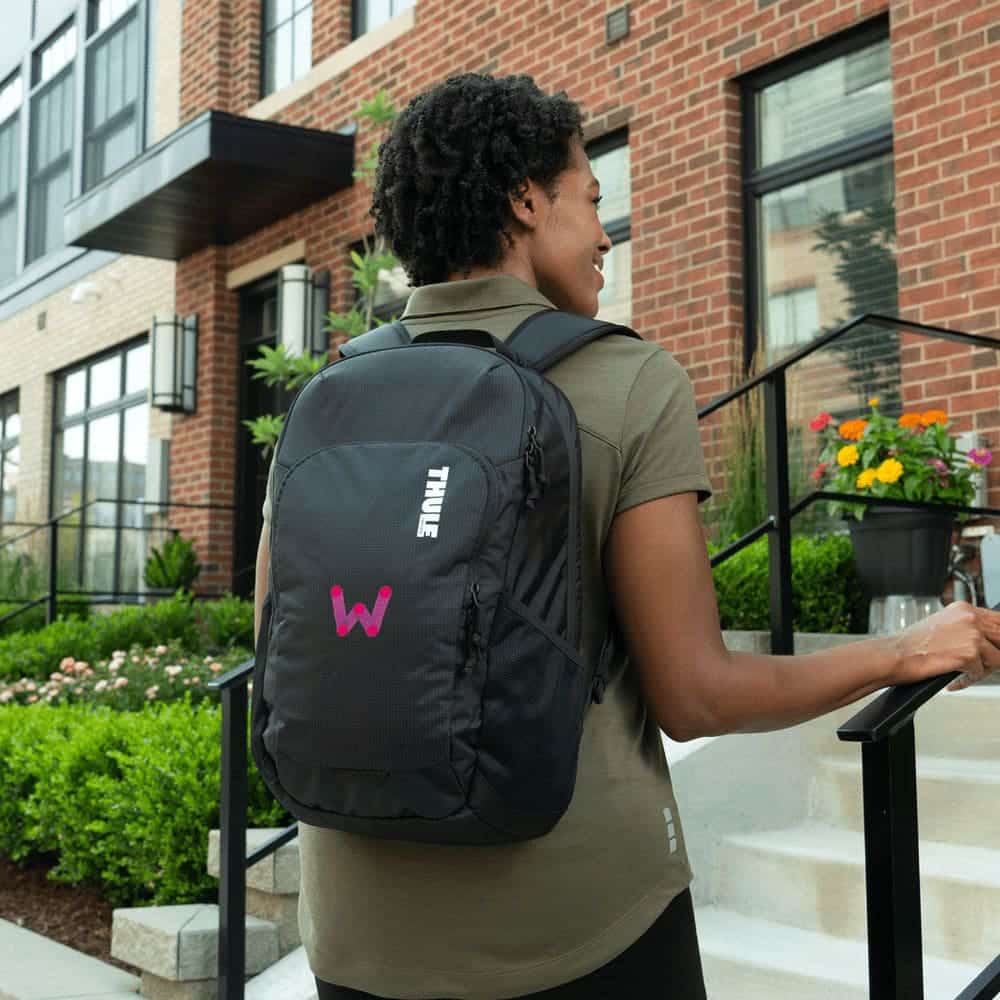 A black Thule Achiever 15 laptop backpack with a logo, worn by a person, in an outdoor urban setting.