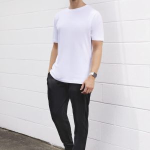 A man stands against a white wall wearing a plain white t-shirt and black track pants, with a watch and sneakers.
