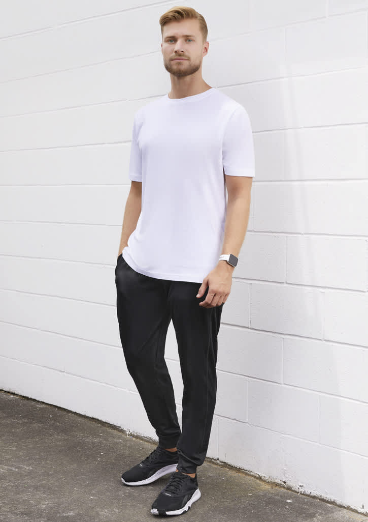 A man stands against a white wall wearing a plain white t-shirt and black track pants, with a watch and sneakers.