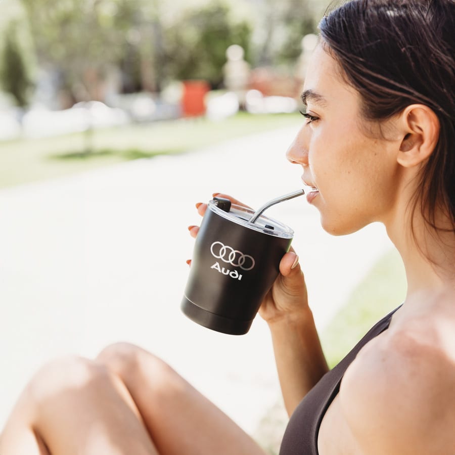 A drink bottle features a matte black design with a straw, held by a person outdoors. It has a logo on it.