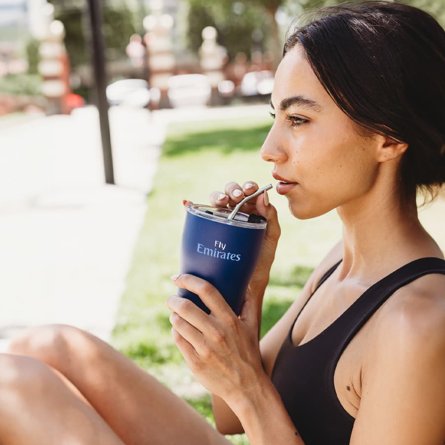 A drink bottle in navy blue with a stainless steel straw and a logo, held by a woman outdoors.