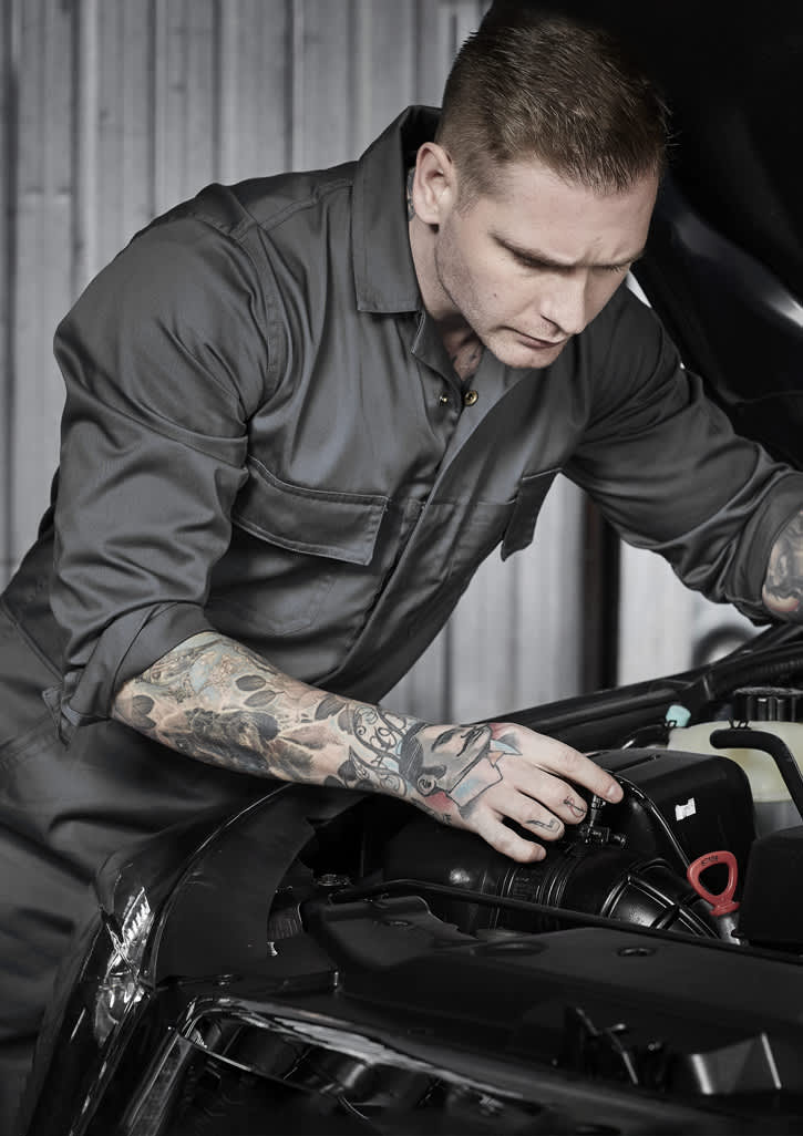 A man wearing a dark grey overall inspects the engine of a car, showcasing detailed tattoos on his arm.