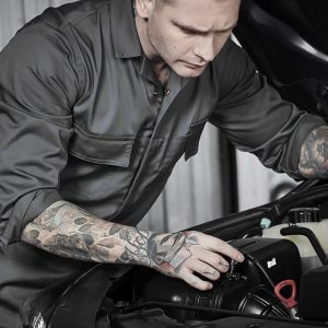 A man in a grey service overall works on a car engine, showcasing detailed tattoos on his arms. The image features a logo on the overall.