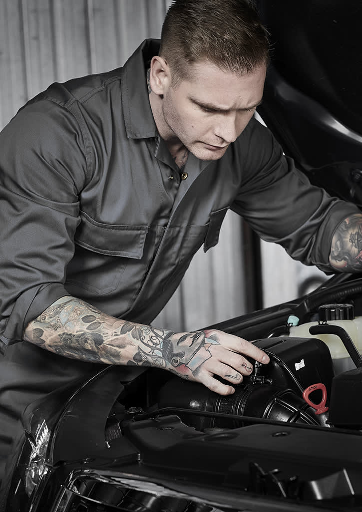 A man in a grey service overall works on a car engine, showcasing detailed tattoos on his arms. The image features a logo on the overall.