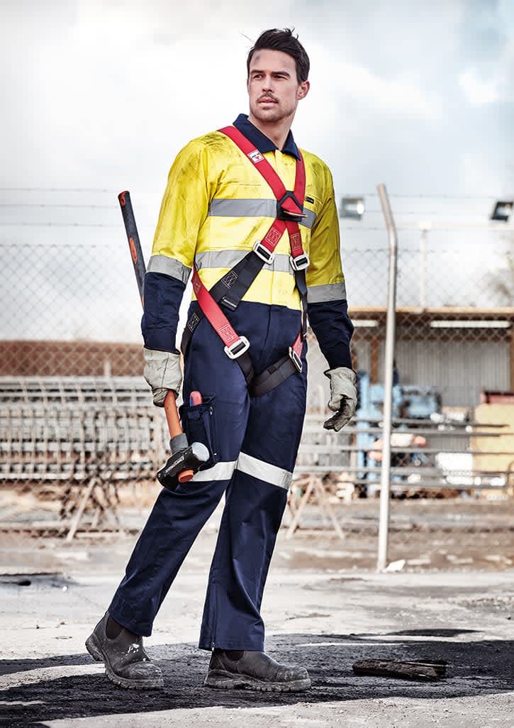 A man in a yellow and navy rugged cooling taped overall with a harness, standing on a construction site.
